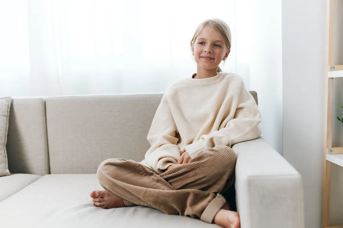 Young girl sits cross-legged on a light sofa, in a sunlit, cozy minimalist living room