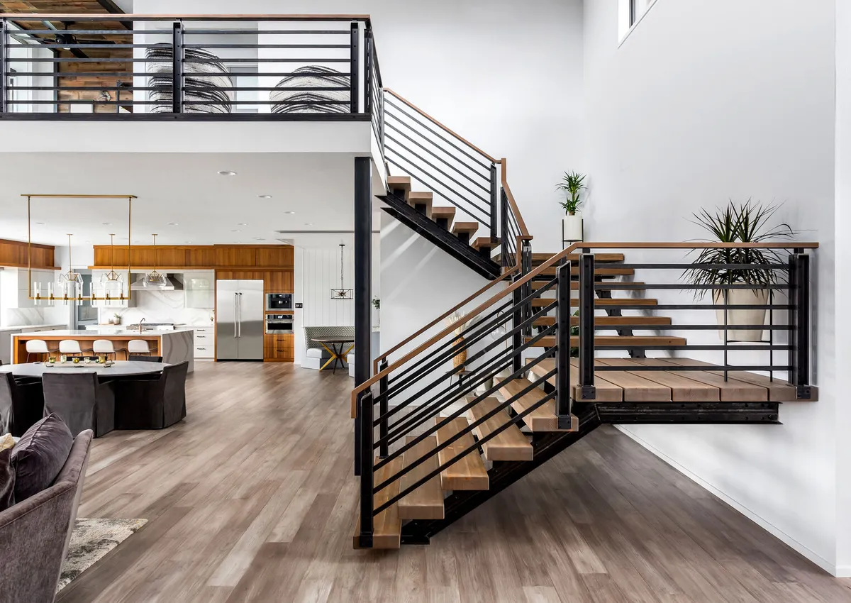 Open concept living room in new luxury home with modern industrial floating staircase and view of kitchen with large island and stainless steel appliances