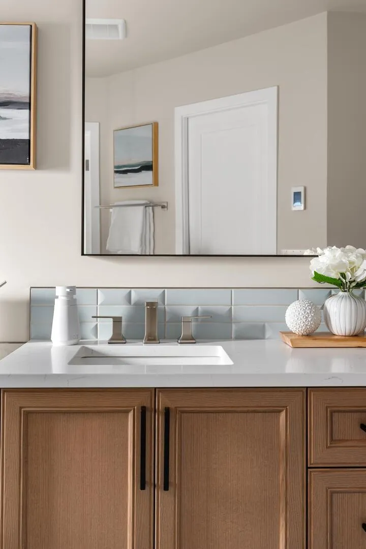 Clean vanity with white counter and wood cabinet with black pulls blue tile backsplash