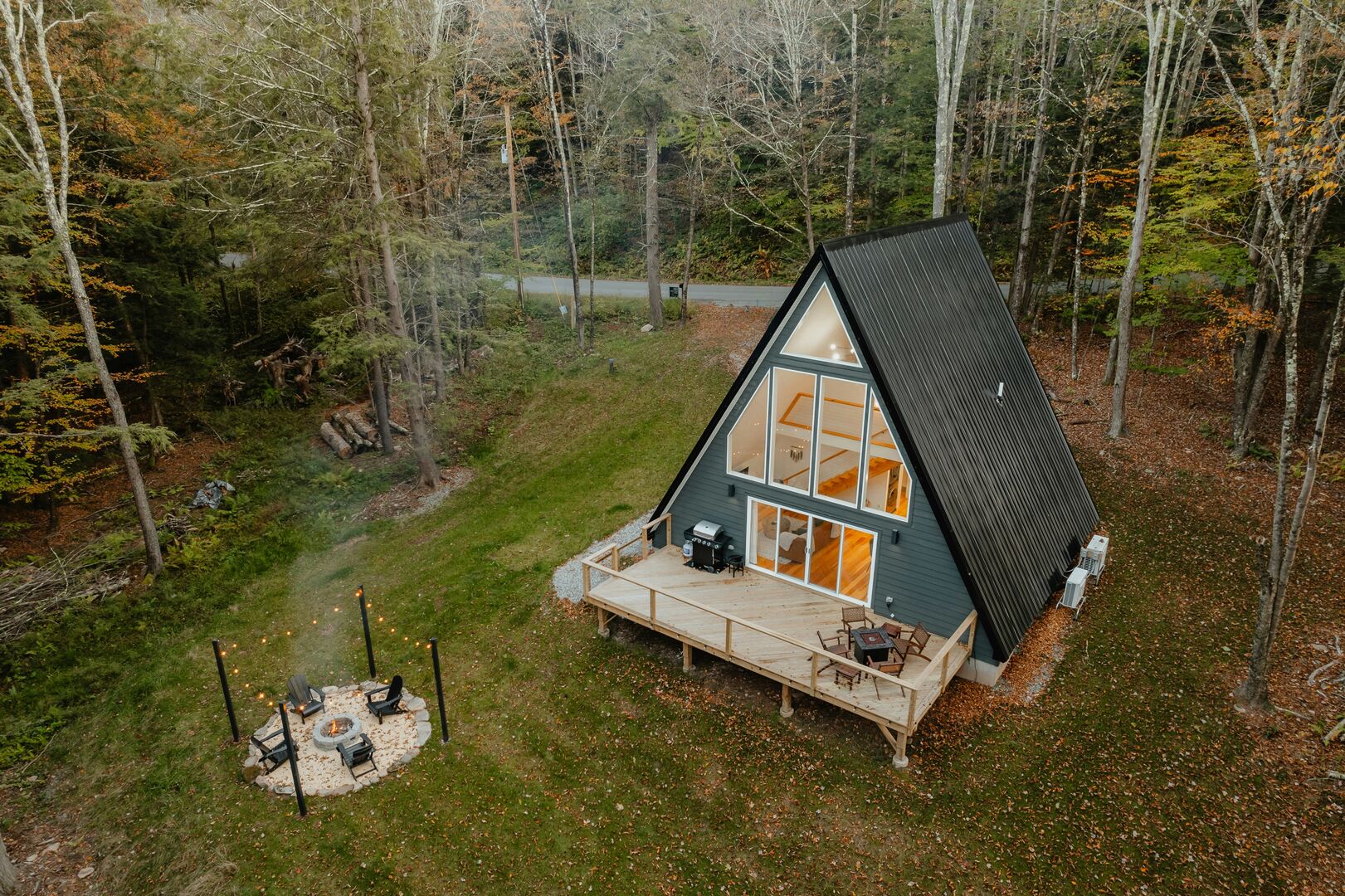 Small a-frame tiny home in the woods with a black metal roof, blue vinyl siding and a wood patio with furniture.