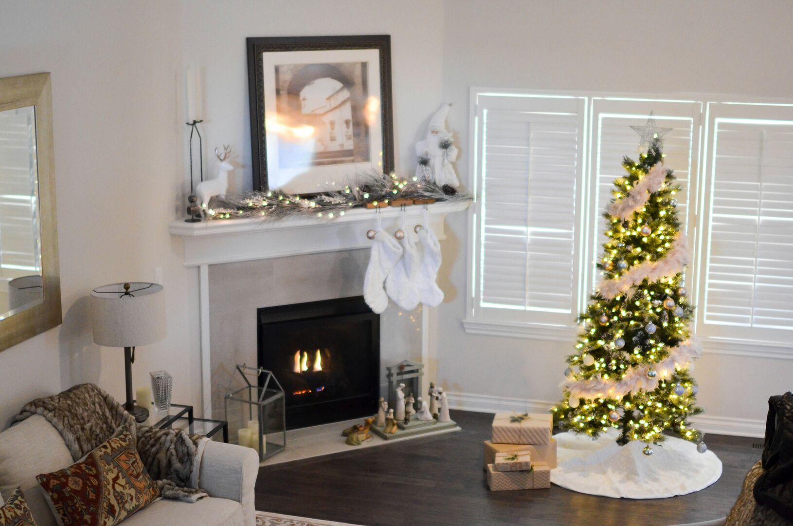 Large living room with a fireplace decorated with a christmas garland and white stockings beside a large christmas tree with white lights.