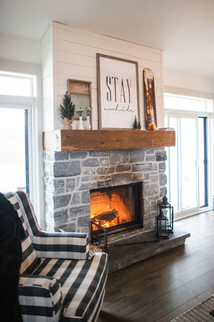 Ski chalet living room with a large brick wood burning fireplace, large windows letting in natural light and a warm wood toned fireplace mantel.