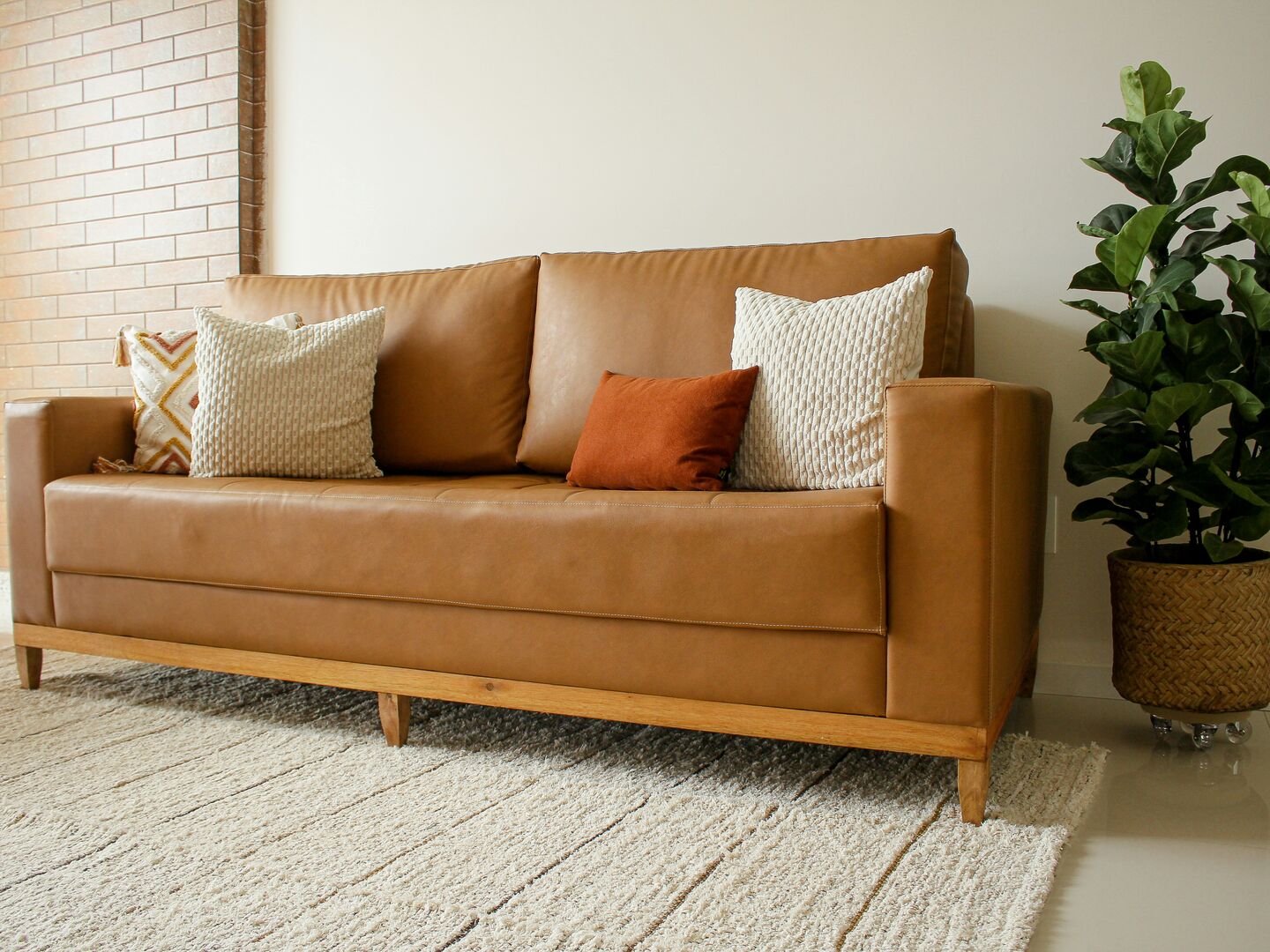Brown leather couch in a living room with rust and cream coloured throw pillows beside a woven potted floor plant.
