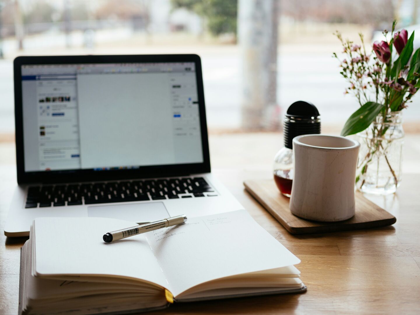 A white and black Macbook laptop on a table with an open notebook and a mug of coffee. 