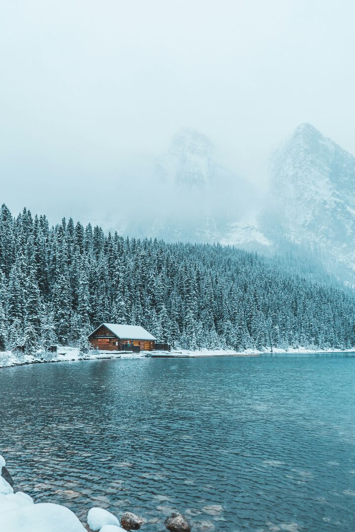 Wood log cabin beside a large blue lake in front of the mountains on a snowy winter day.