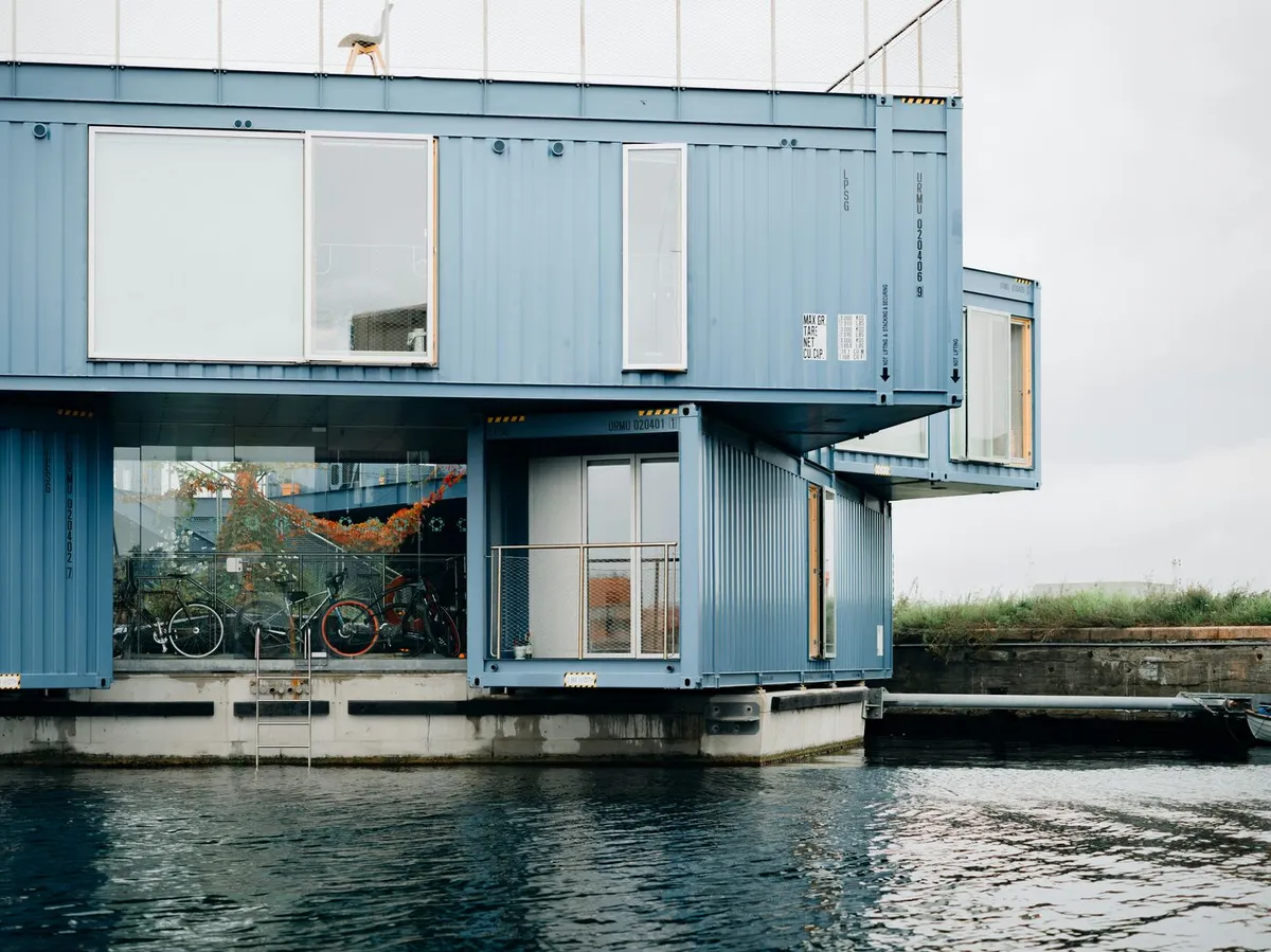 A modern blue shipping container tiny home with large windows on a riverfront property